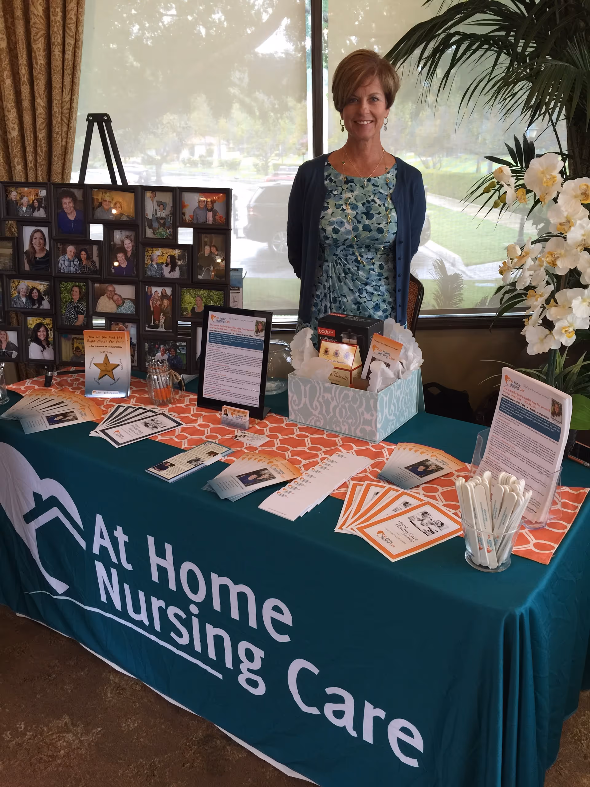 Smiling woman stands at indoor promotional table for At Home Nursing Care.