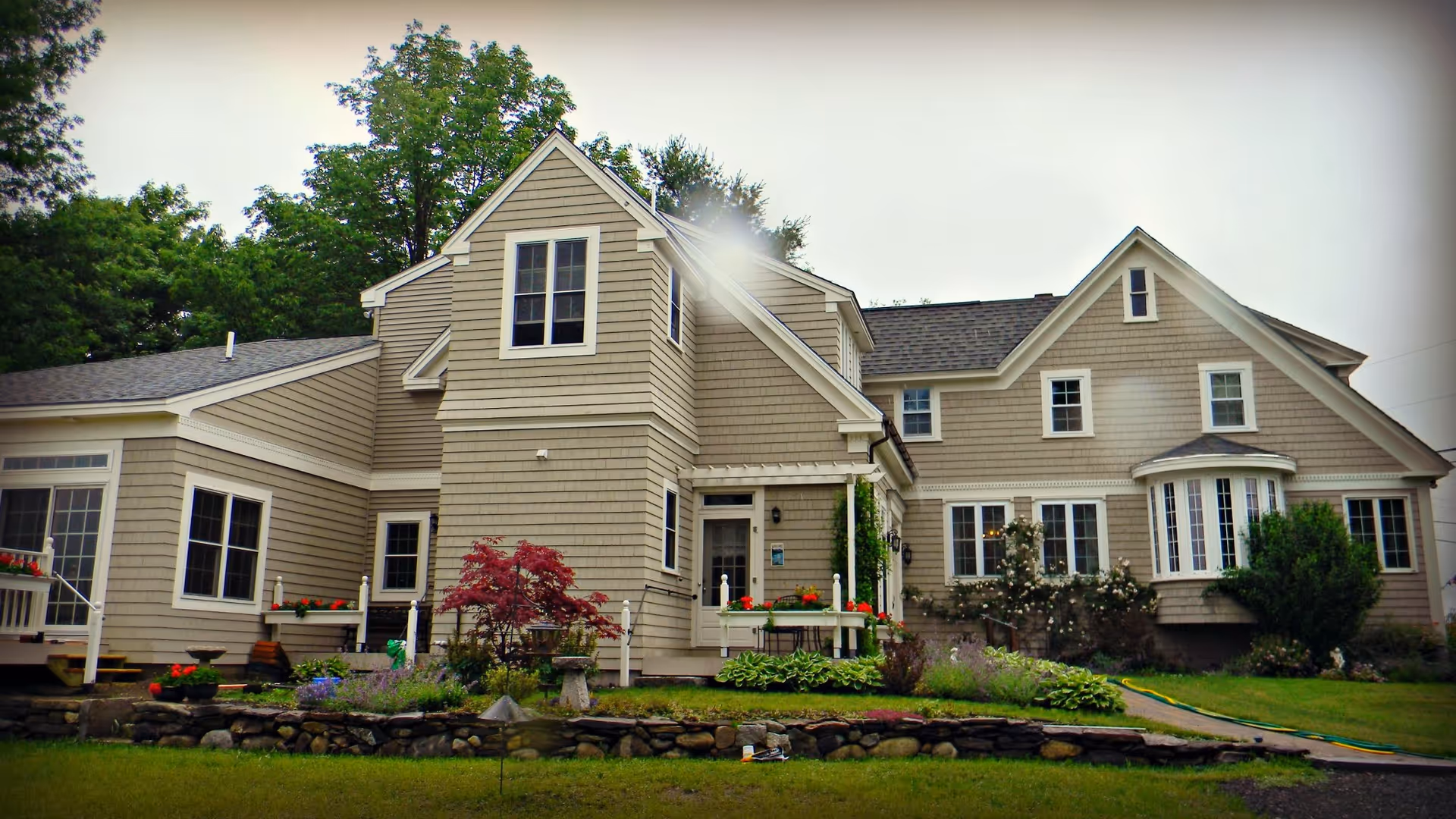 Exterior view of a large beige two-story house with multiple windows, a small porch, and a well-maintained garden with various plants and flowers in front. The sky is overcast.