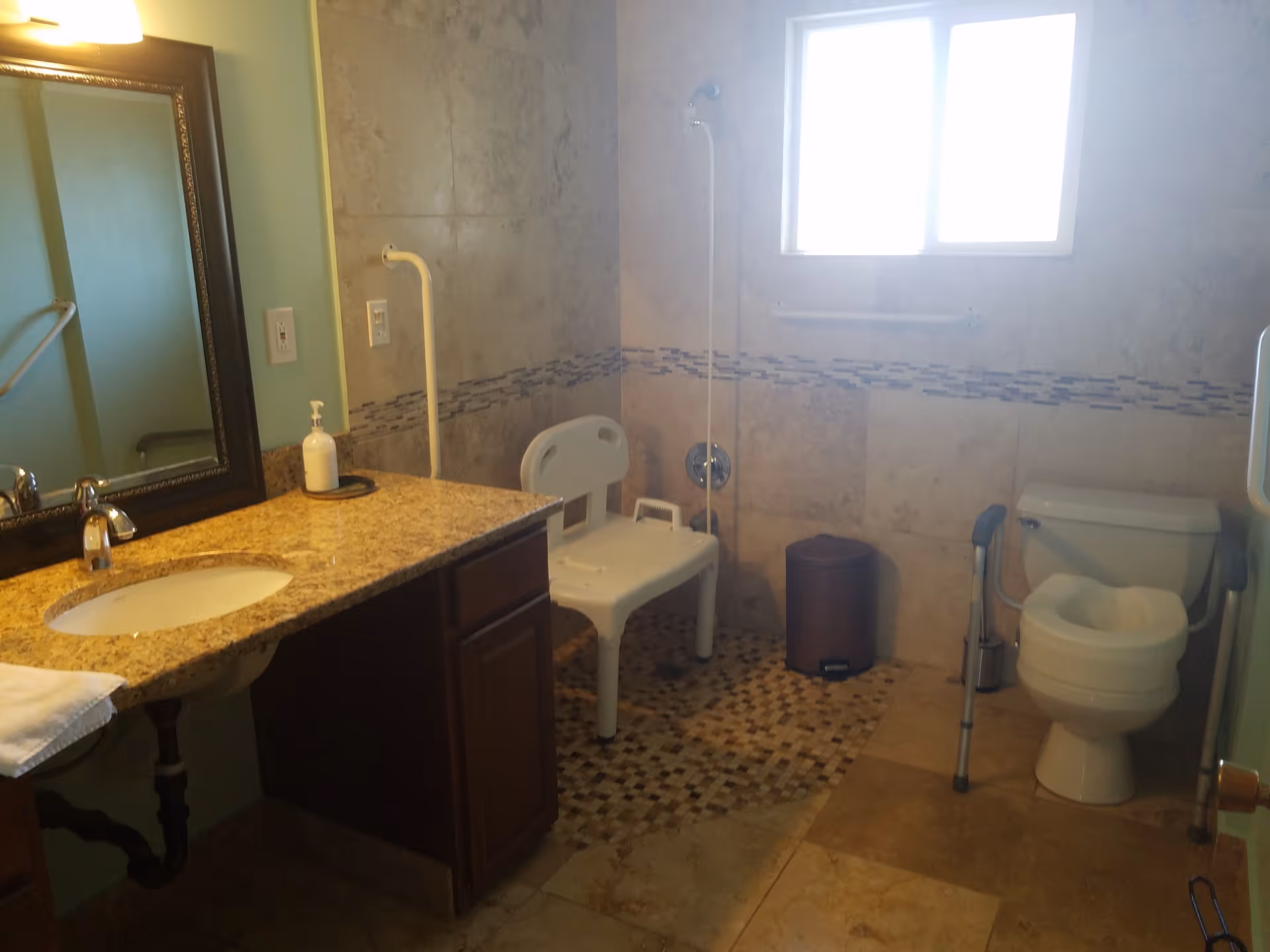 Accessible bathroom with sink, mirror, shower chair, grab bars, toilet, and tiled walls.
