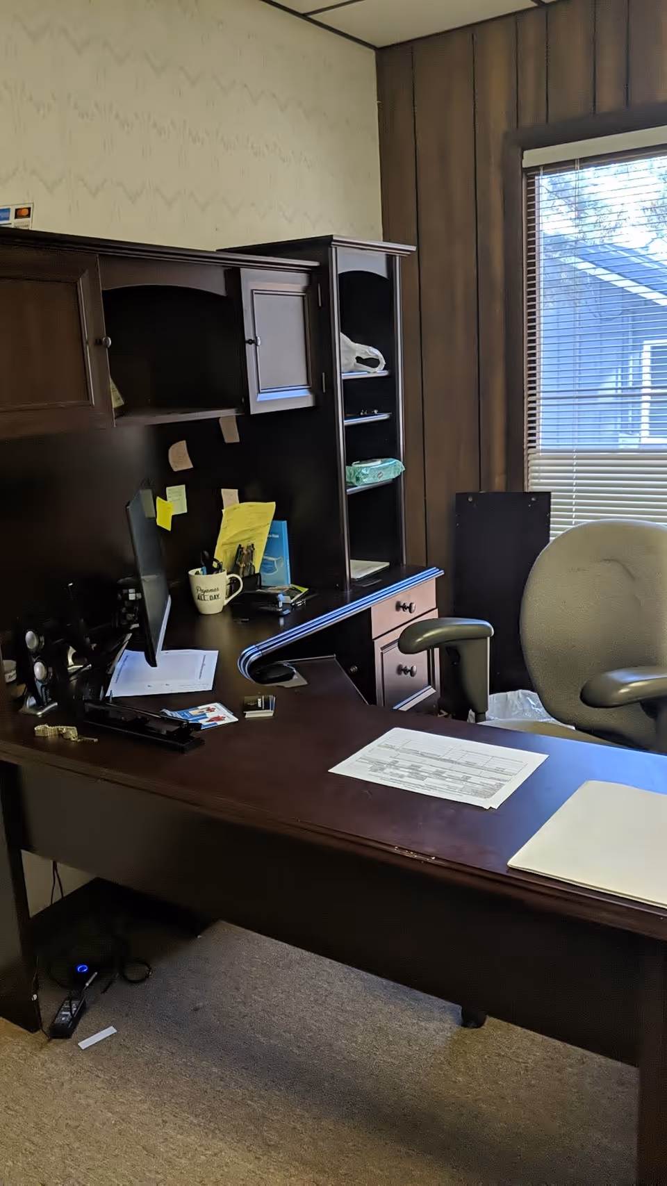 Dark wooden desk with computer and shelving occupies a small, windowed office space.