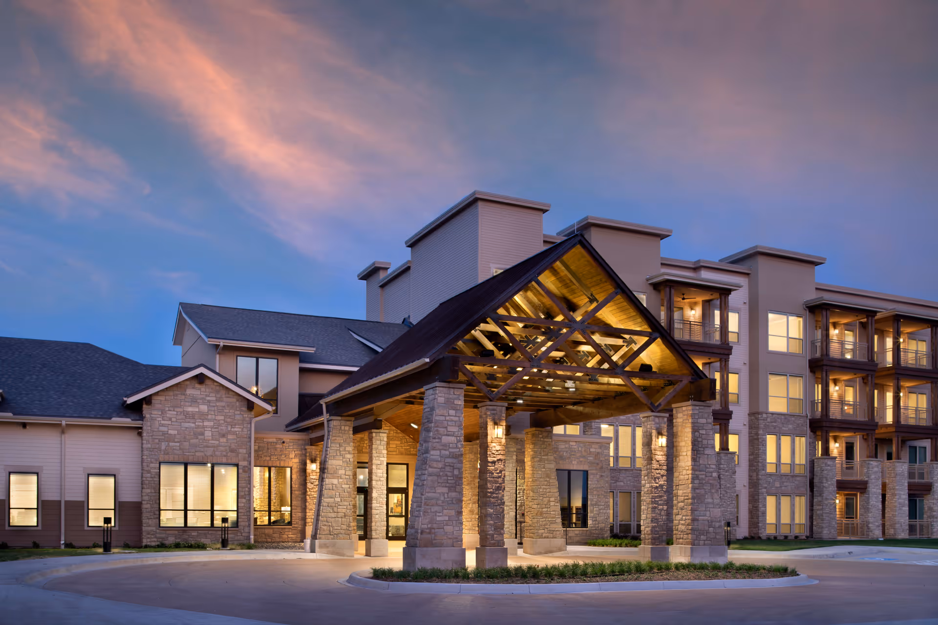 Exterior view of Stevenson Oaks facility at dusk, featuring a large covered entrance with stone pillars and warm lighting, multi-story building with balconies and large windows under a partly cloudy sky.