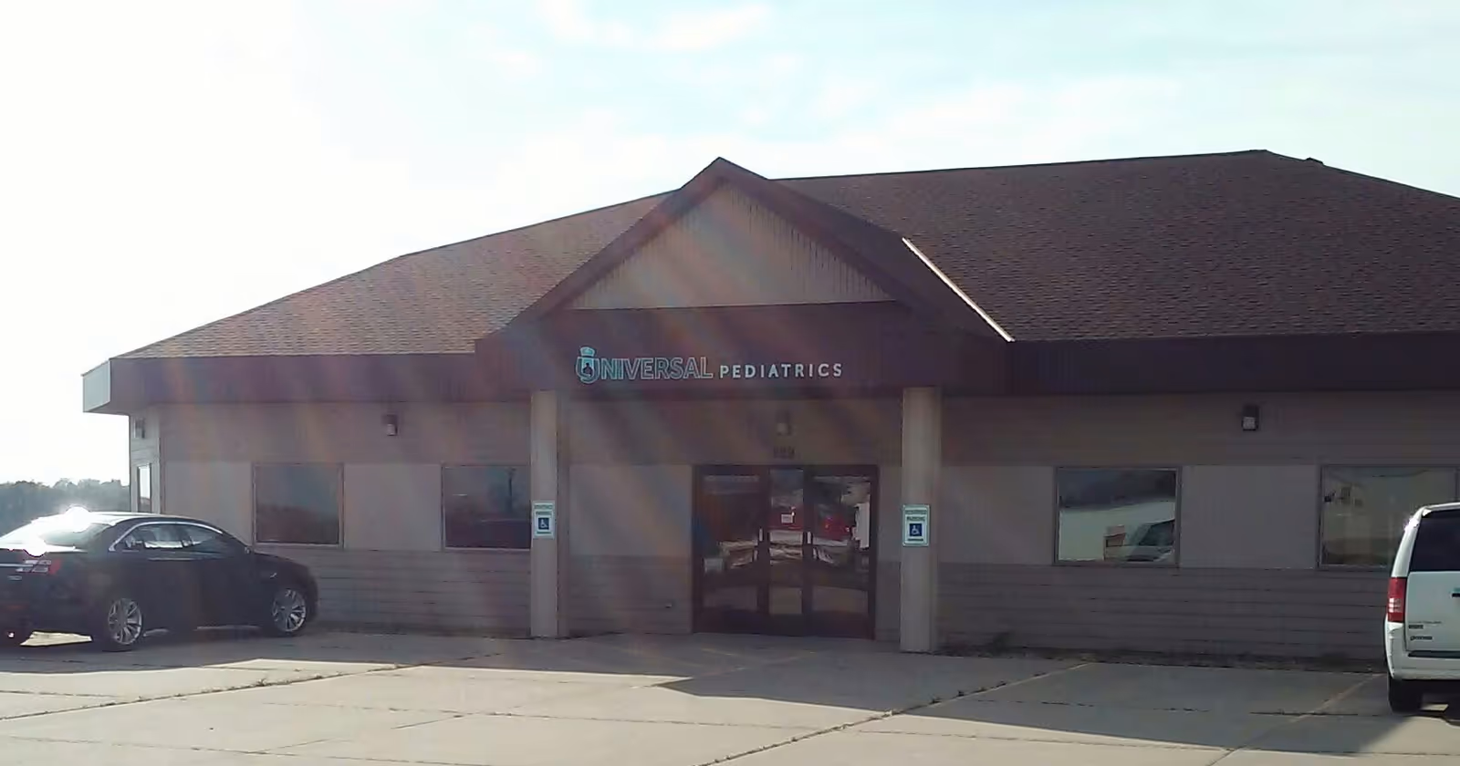 Single-story beige building with glass entrance labeled Universal Pediatrics and parked cars.