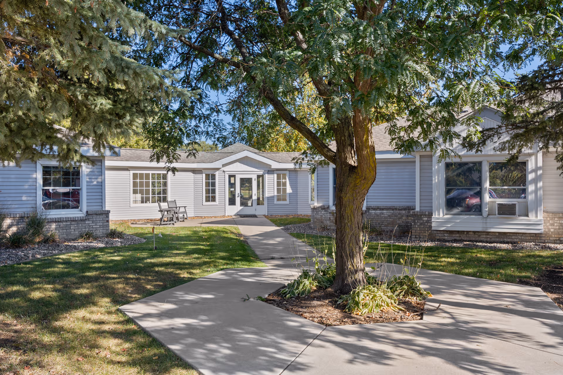 Front exterior of a single-story senior living facility with a central tree and walkway.