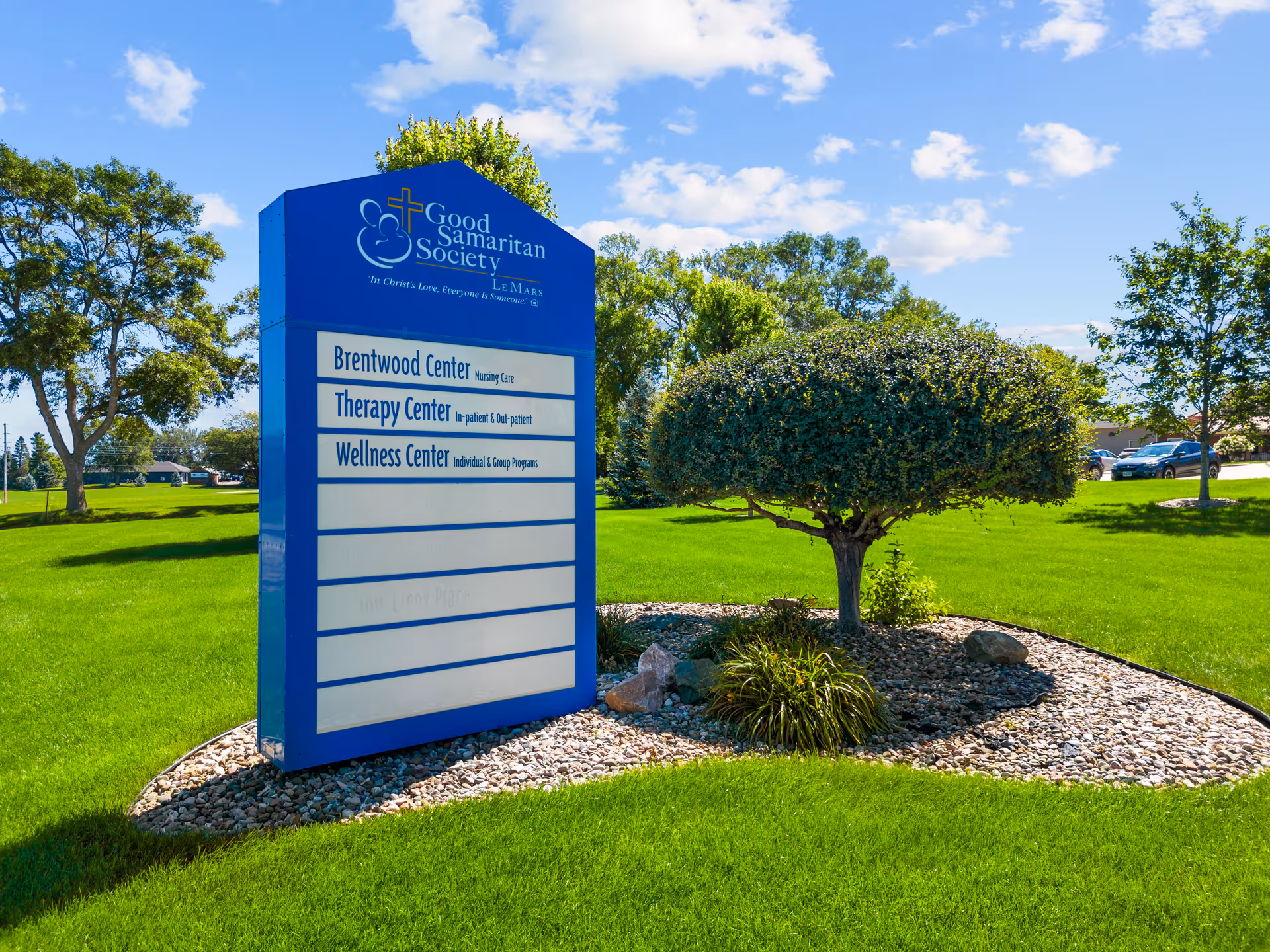 Outdoor view of a blue sign for Good Samaritan Society - Le Mars, listing Brentwood Center Nursing Care, Therapy Center In-patient & Out-patient, and Wellness Center Individual & Group Programs, surrounded by green grass, trees, and a small landscaped area with rocks and bushes under a partly cloudy sky.