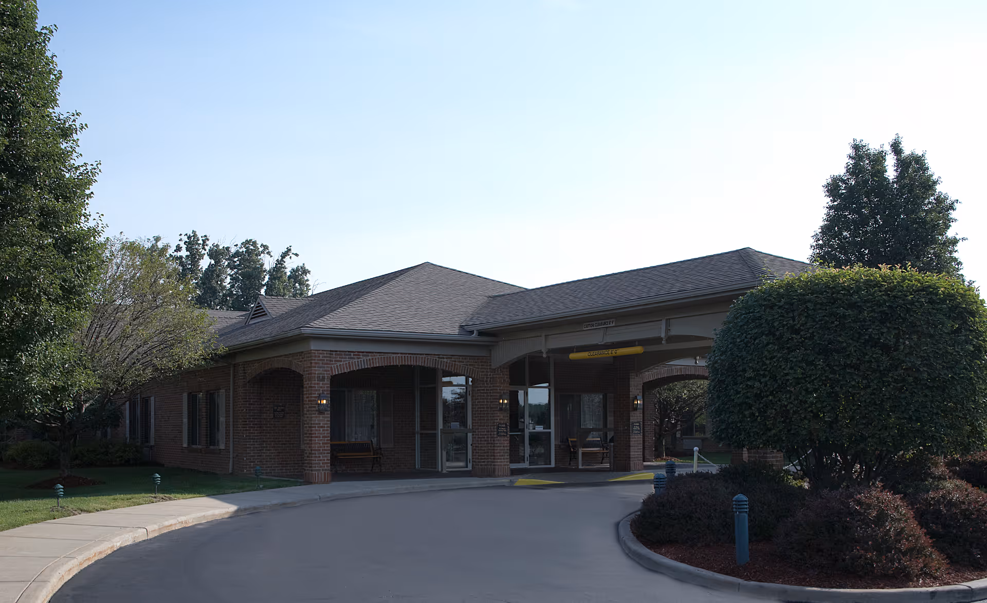 Brick one-story senior living building entrance with a covered porte-cochere, circular driveway, and landscaped trees and shrubs.