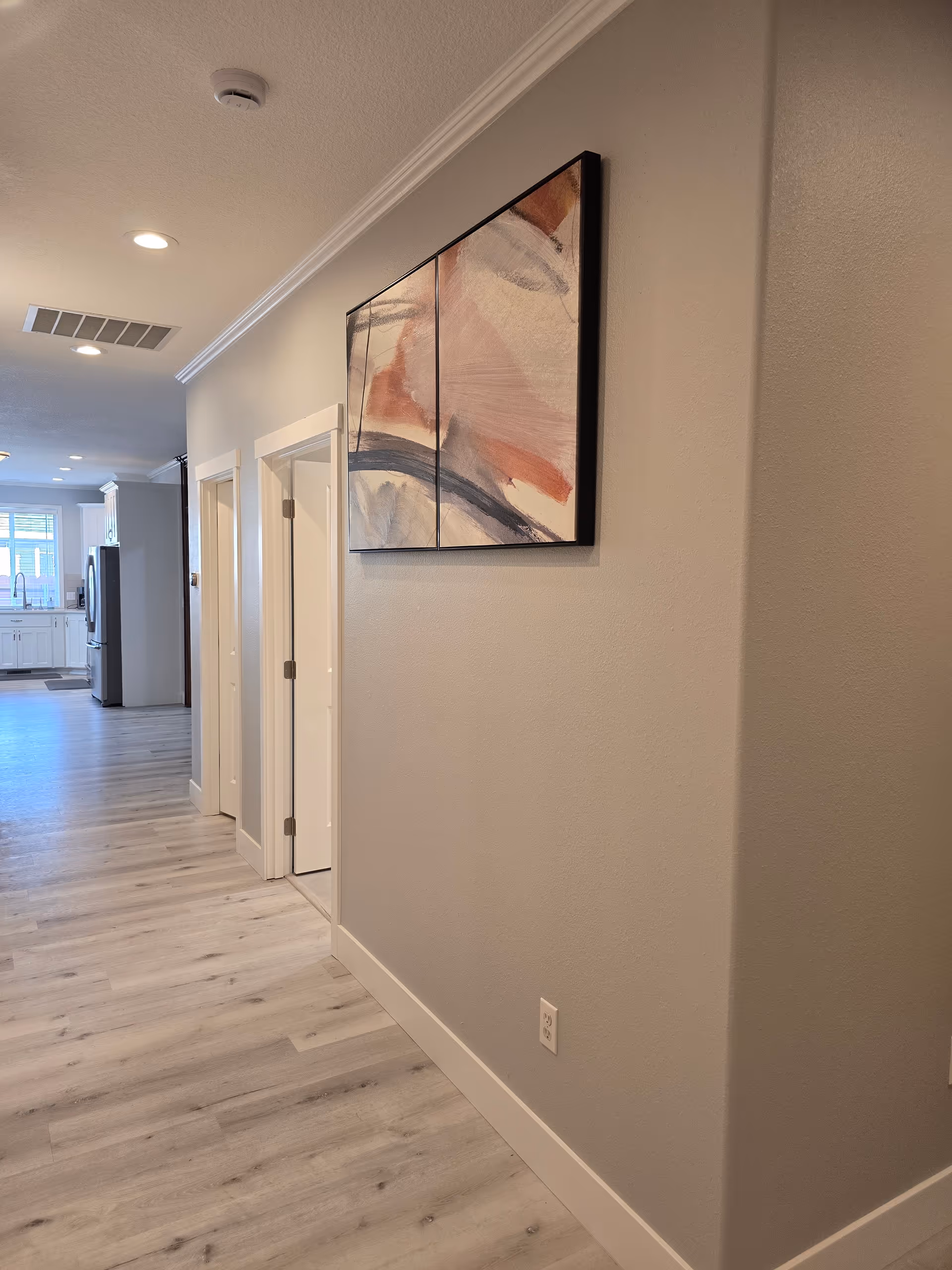 Bright hallway inside Sunquest Care Home, LLC showing wall art, doors, and a distant kitchen.