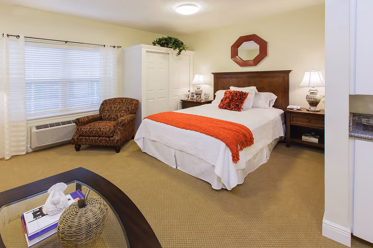 A cozy bedroom with a neatly made bed featuring white bedding and a red throw blanket and pillow. There are two wooden nightstands with lamps on either side of the bed, a decorative red mirror above the headboard, a patterned armchair near a window with white curtains, and a white wardrobe. A glass coffee table with decorative items is partially visible in the foreground.