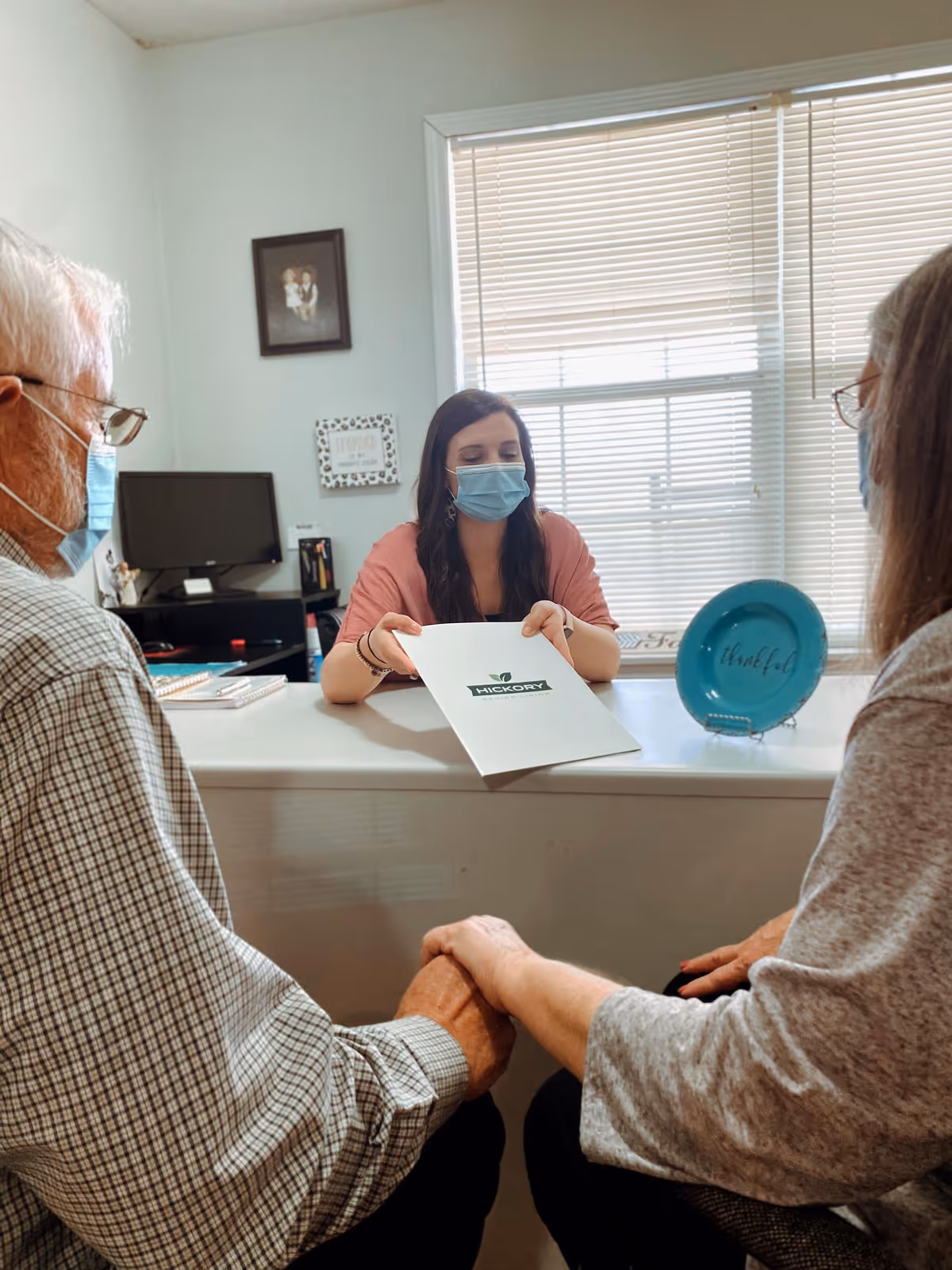 A woman wearing a face mask sits behind a desk holding a folder labeled 'Hickory Senior Living' while talking to an elderly couple who are holding hands. The room has a window with blinds, a computer monitor, and framed pictures on the wall.