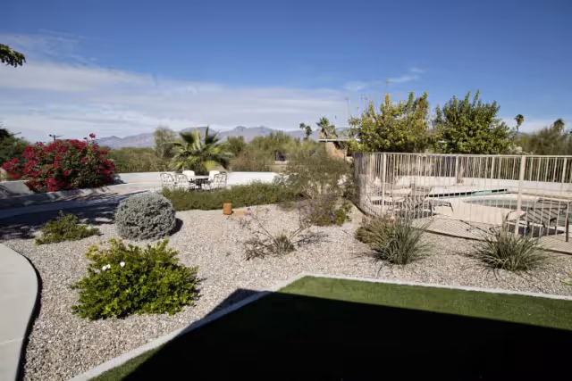 Desert landscaped backyard with gravel, shrubs, a fenced pool, patio seating, and distant mountains under a blue sky.