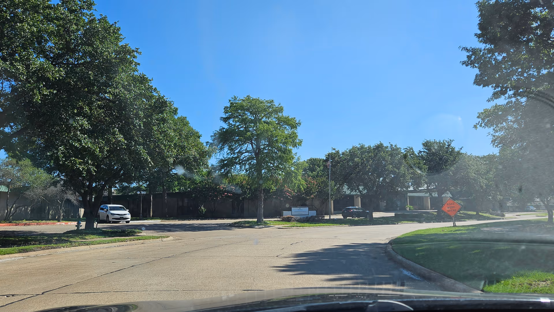 Curved road leads to a facility surrounded by trees with parked cars and a construction sign.