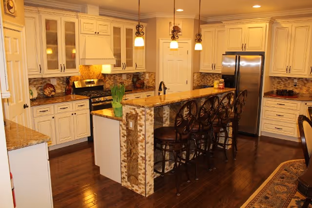 A spacious kitchen with cream-colored cabinets, a large island with a granite countertop, and four high-back bar stools. The kitchen features stainless steel appliances including a refrigerator and a stove with a range hood. Pendant lights hang above the island, and the floor is made of dark hardwood. The backsplash has a mosaic tile design, and there is a potted plant on the island.