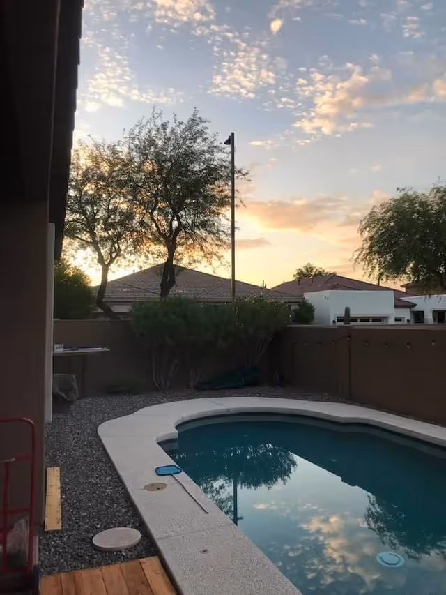 Outdoor pool in a fenced backyard with desert landscaping and sunset sky.