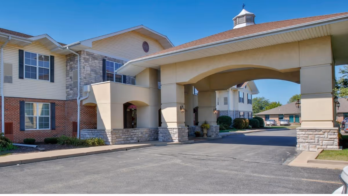 Exterior view of a senior living facility with a covered entrance supported by large columns. The building features a combination of brick, stone, and siding with multiple windows. There is a paved driveway and some landscaping with bushes and grass.