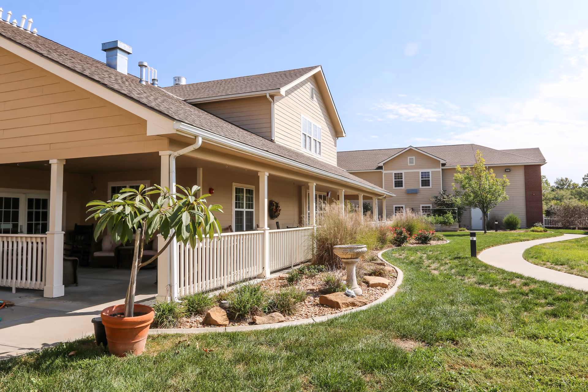 Front exterior of a beige senior living building with a covered porch, landscaped lawn, and a curved sidewalk.