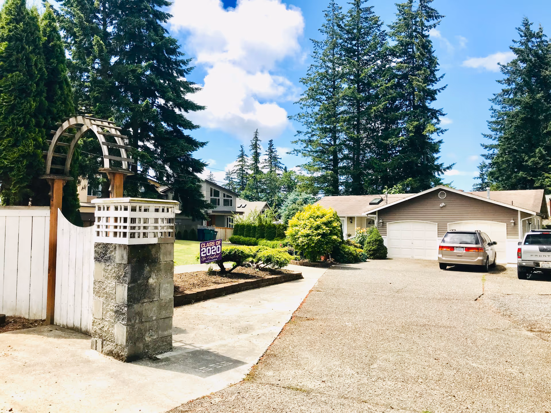 Senior living facility exterior with driveway, garage, cars, and tall trees.