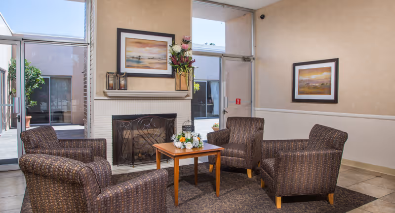 A cozy seating area with four patterned armchairs arranged around a wooden coffee table with a floral centerpiece. The room features a white brick fireplace with a decorative screen, a framed landscape painting above the mantel, and a tall vase with flowers. Large windows and glass doors provide a view of an outdoor courtyard with potted plants.