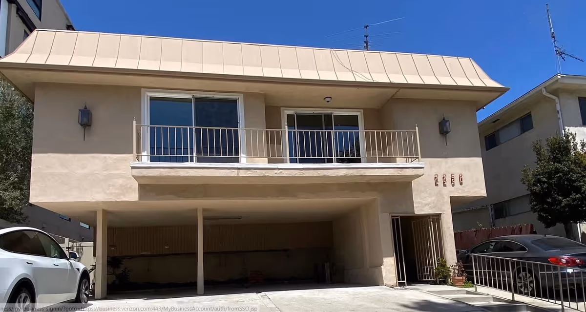 Two-story beige building with balcony and open carport, with parked cars.