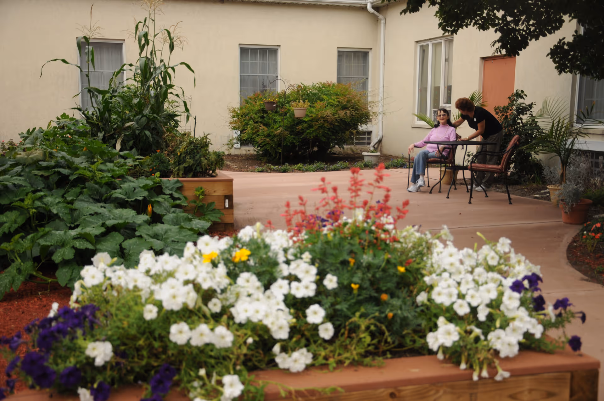 Outdoor garden area with colorful flowers in the foreground and two women interacting near a table and chairs on a paved patio in front of a beige building.