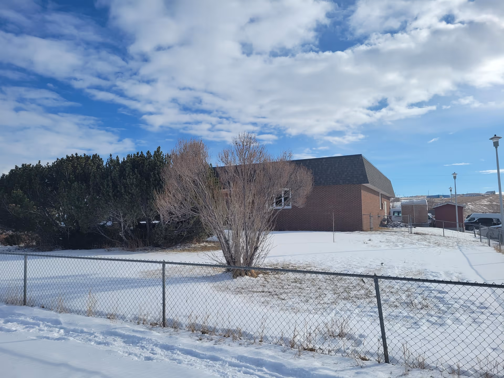 Snowy front yard of a brick senior living facility with a chain-link fence and blue sky.