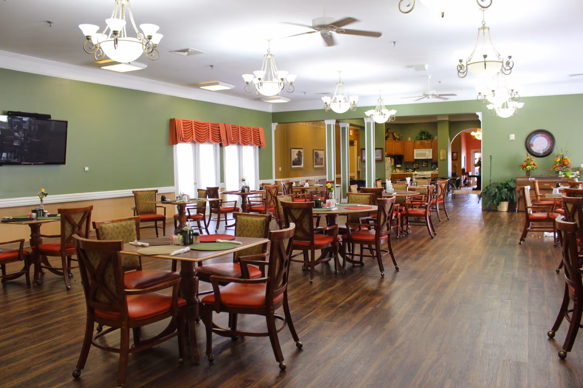 Spacious senior dining room with multiple round tables and chairs, green walls, chandeliers, and wood flooring.