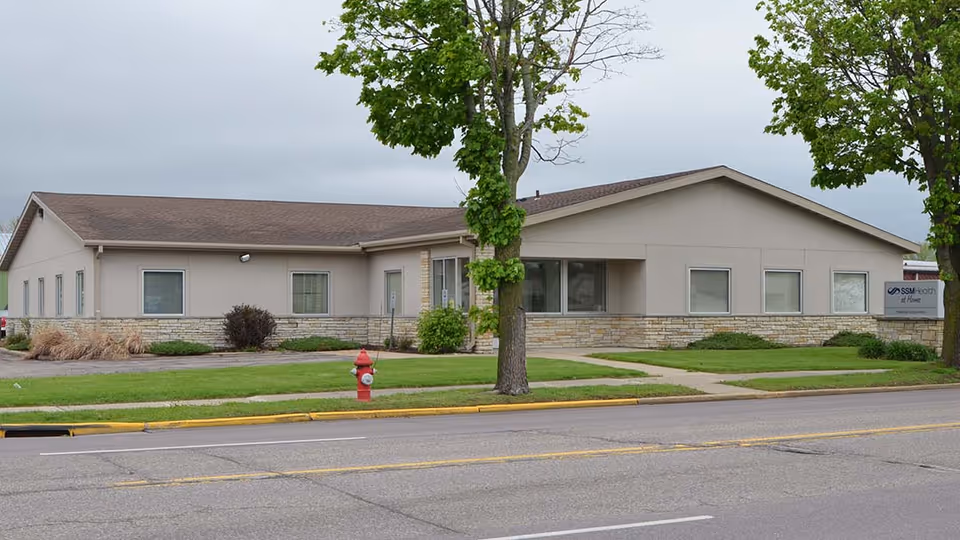 A single-story beige facility building with SSM Health at Home sign along a tree-lined street.