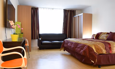 A cozy hotel-style bedroom with a bed, sofa, wardrobe, desk, and large window.