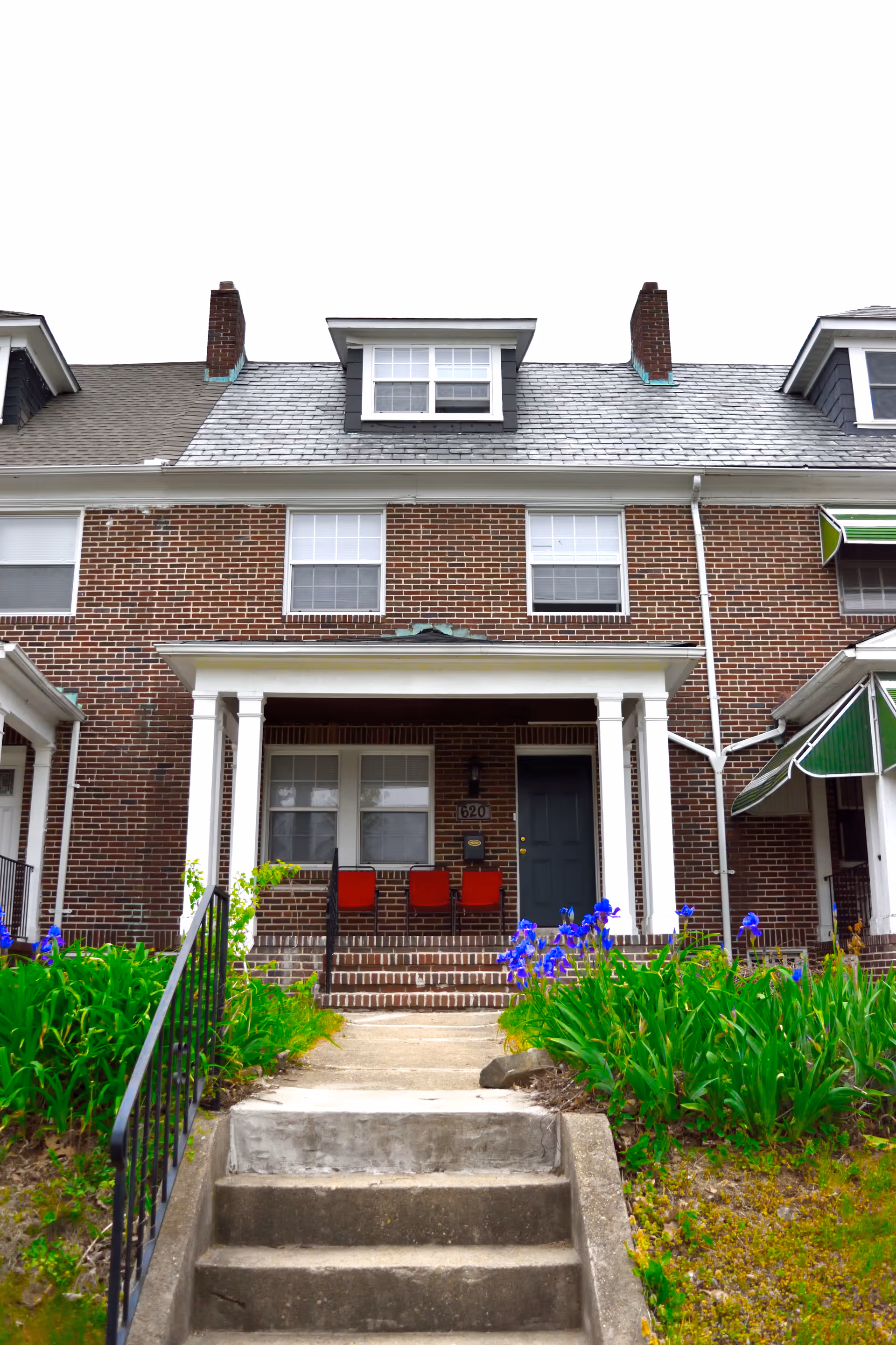 Brick senior living building exterior with a porch, steps, red chairs, and blooming purple flowers.