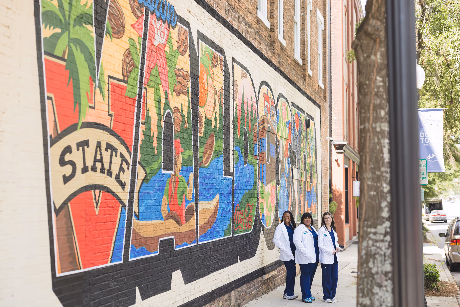 Three caregivers stand beside a colorful mural on a sidewalk outside a building.