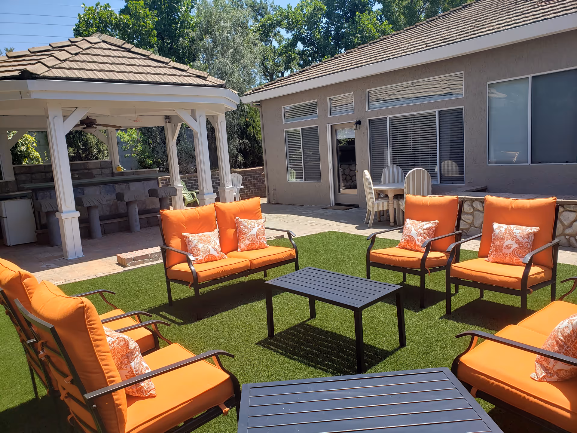 Outdoor patio with orange-cushioned chairs and loveseats arranged around low tables on artificial grass beside a gazebo and building.