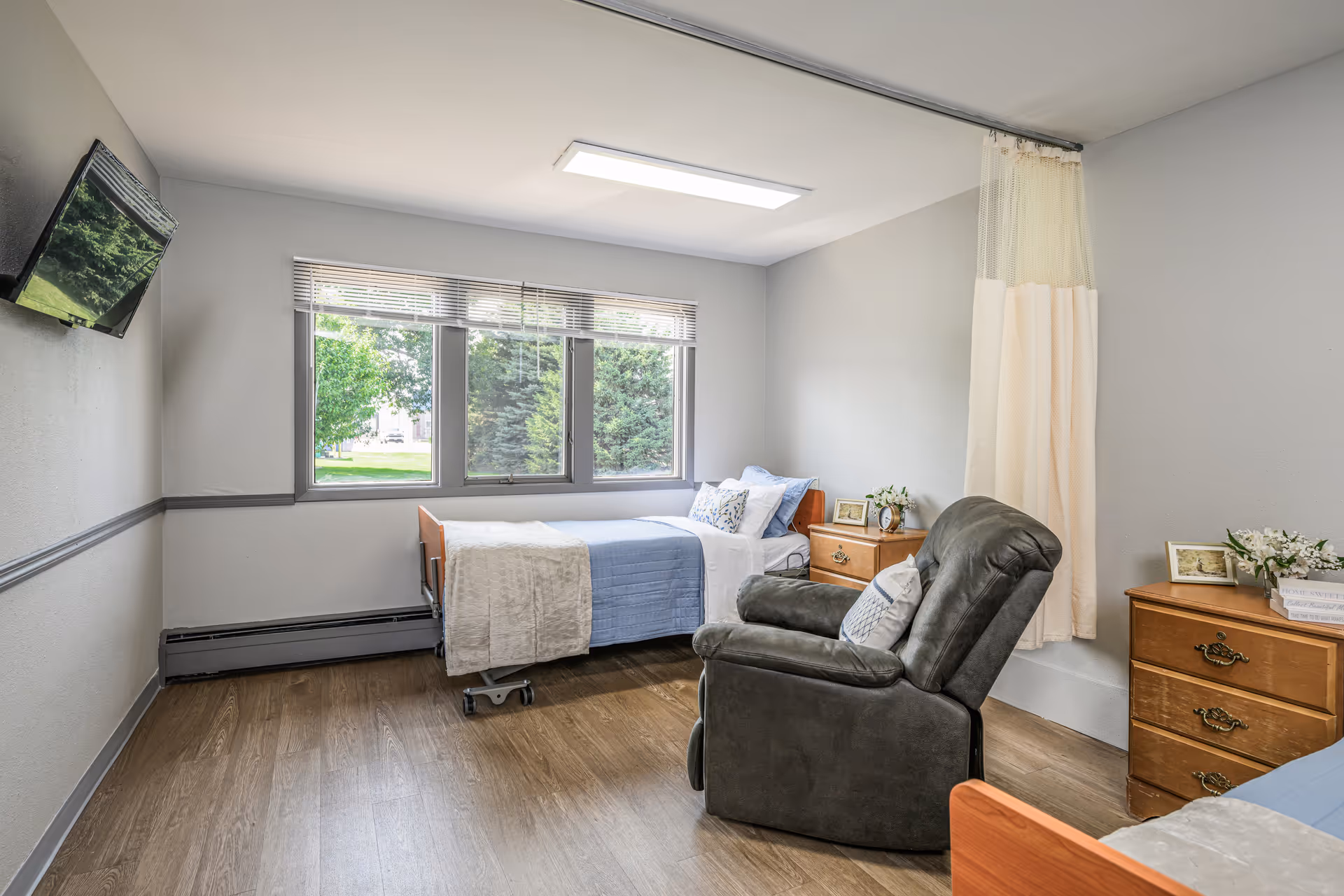 A furnished senior living bedroom with bed, recliner, nightstands, and a window view.