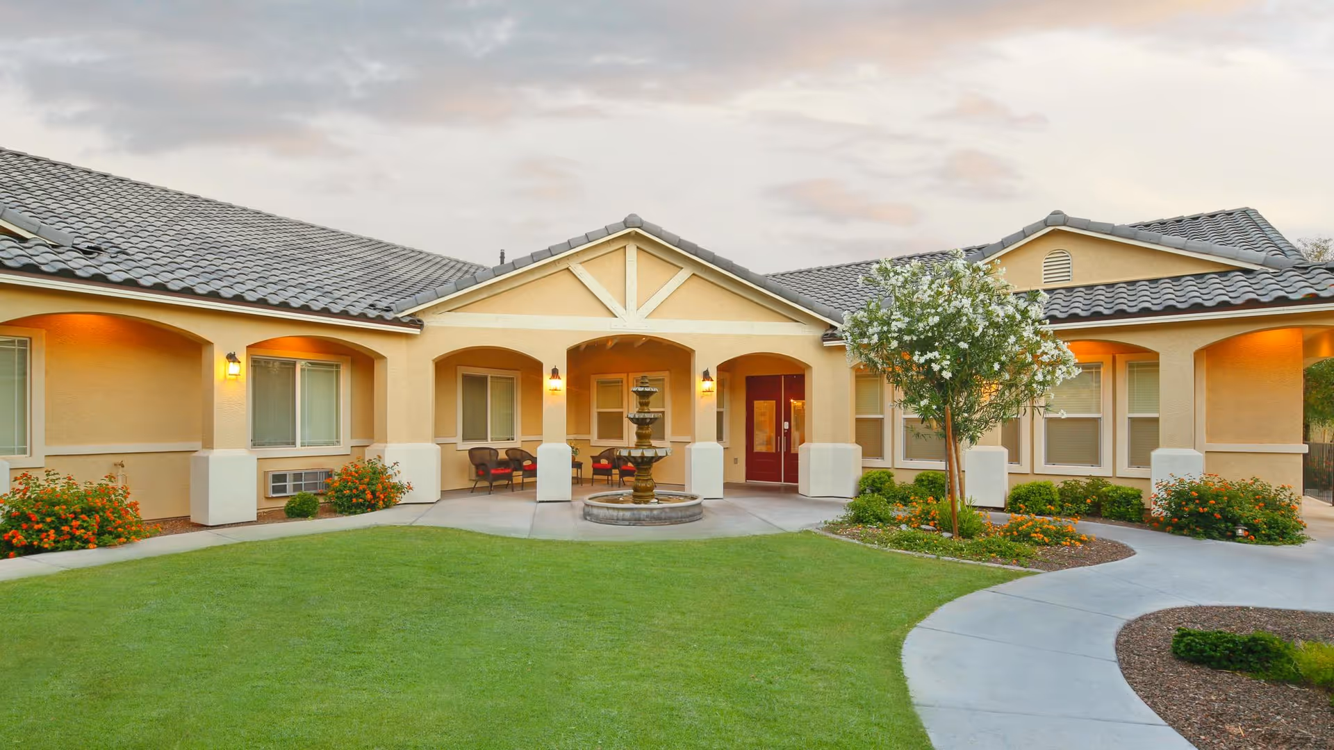 Front entrance of a single-story assisted living facility with a central fountain, lawn, and covered walkway.