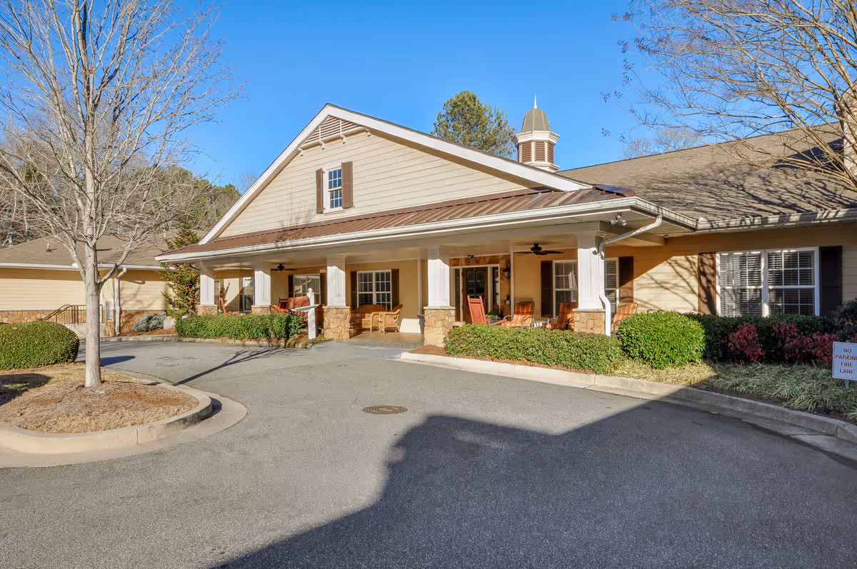 Exterior view of a senior living facility building with a covered porch featuring several chairs. The building has beige siding, stone accents on the porch columns, and a small cupola on the roof. There are some bushes and a tree in the landscaped area near the driveway under a clear blue sky.