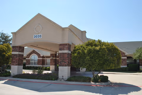 Exterior view of a senior living facility building with a covered entrance supported by brick and beige columns. The building number 3020 is displayed on the front of the entrance. There are trimmed bushes and a tree near the entrance, and the sky is clear and blue.