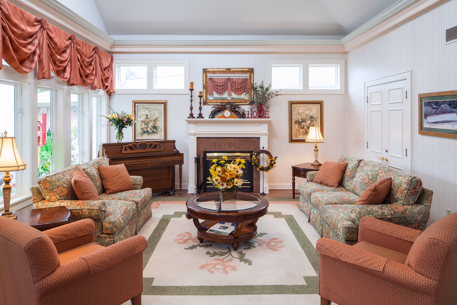 Bright furnished living room with floral sofas and armchairs arranged around a central glass coffee table, a piano, and a fireplace.