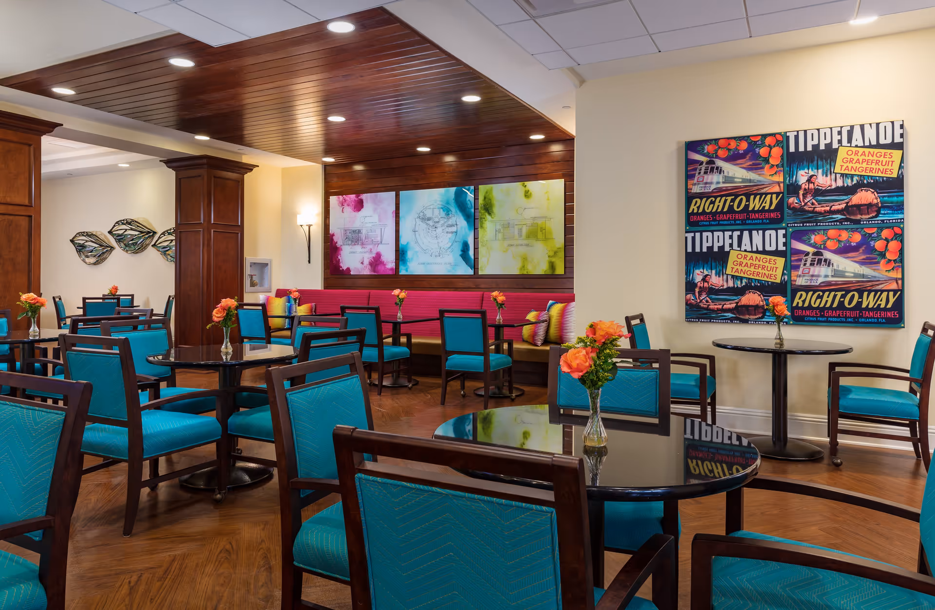 A bright and colorful dining area with multiple round tables and teal upholstered chairs. Each table has a small vase with orange flowers. The room features wooden flooring, a wooden ceiling panel with recessed lighting, and decorative wall art including colorful abstract paintings and vintage-style posters advertising Tippecanoe oranges, grapefruit, and tangerines.