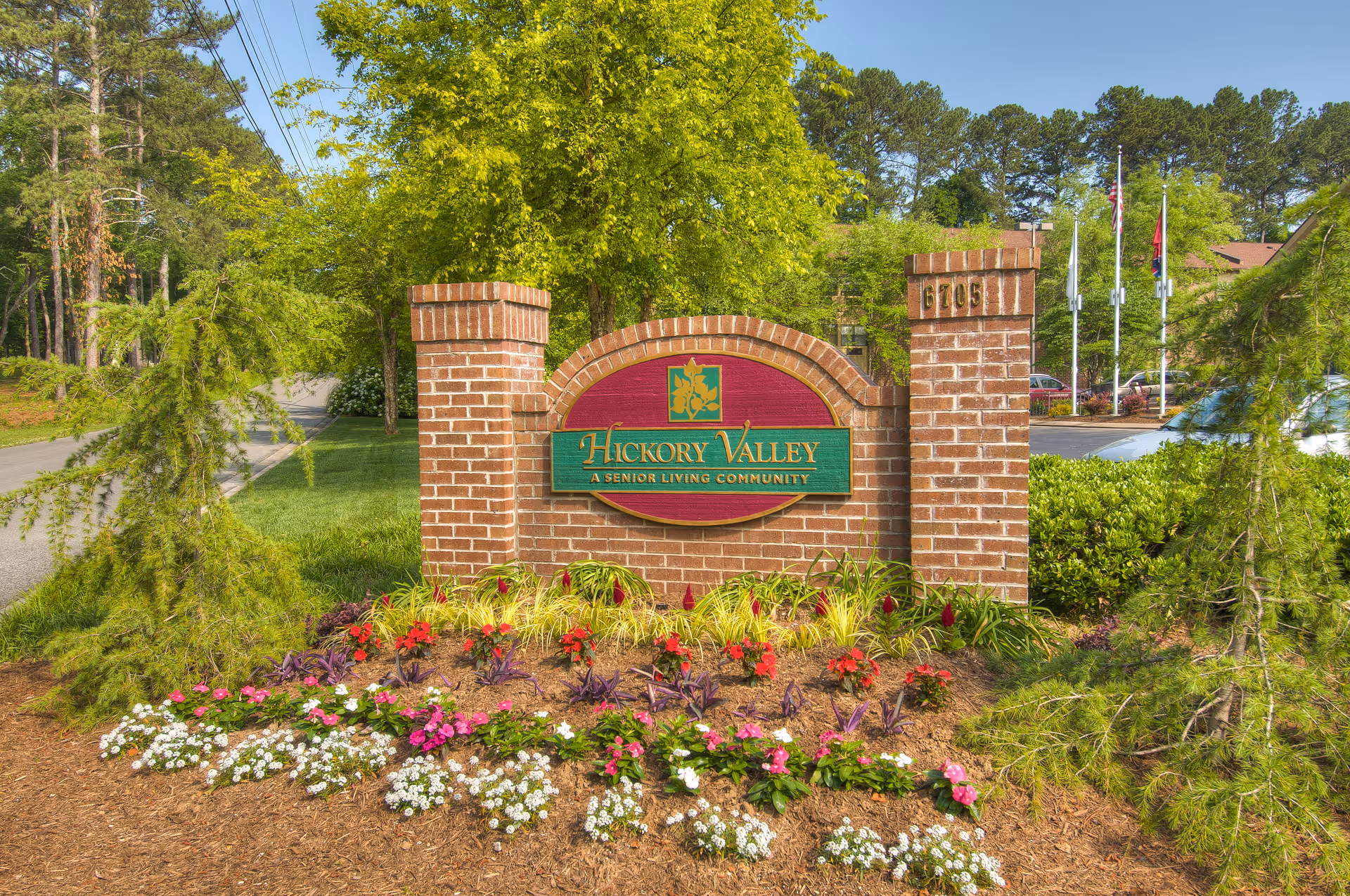 Brick entrance sign for Hickory Valley A Senior Living Community surrounded by colorful flowers and greenery with trees and a road in the background.