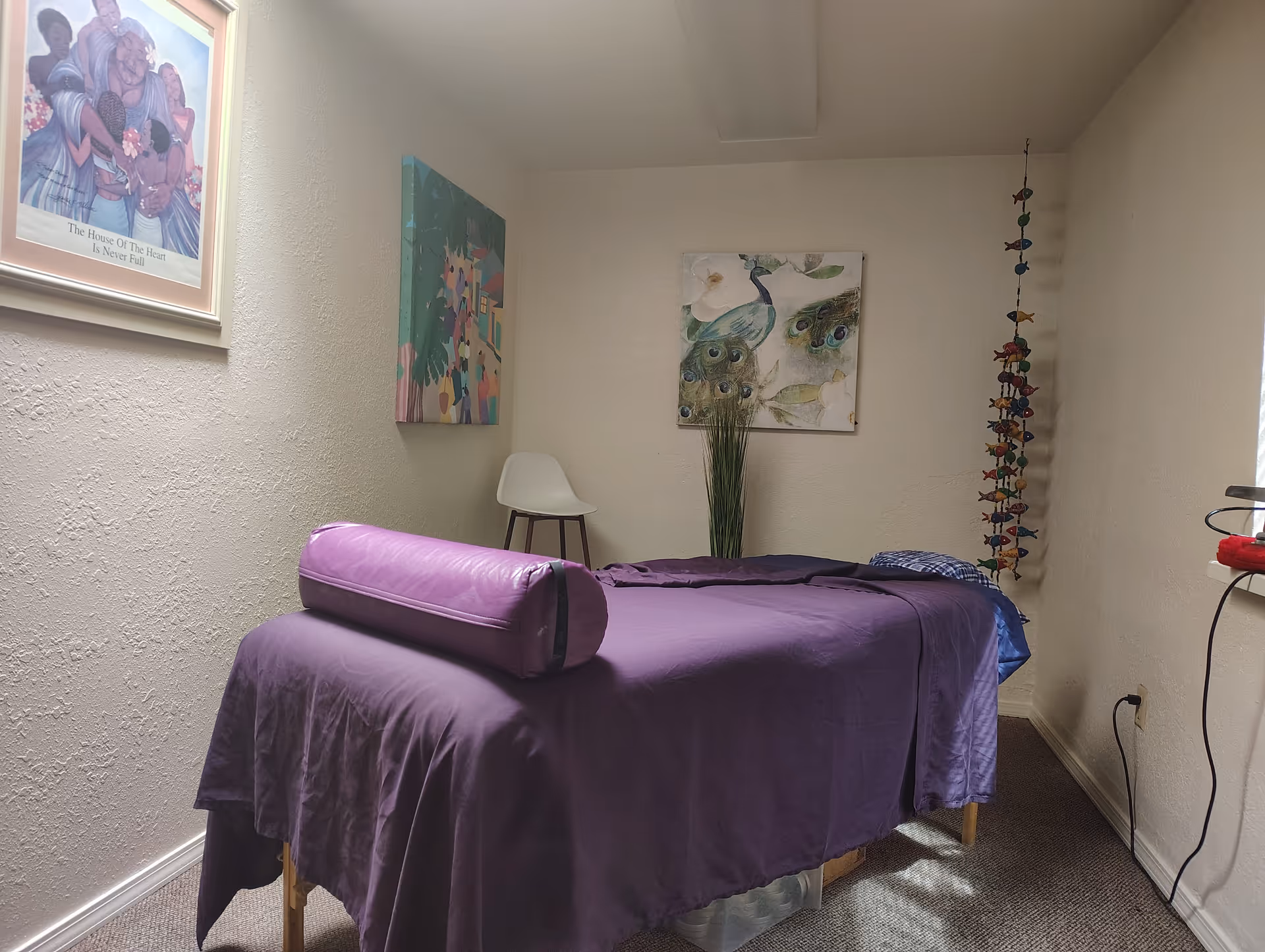 Treatment room featuring a purple-covered massage table, wall art, and a white chair.
