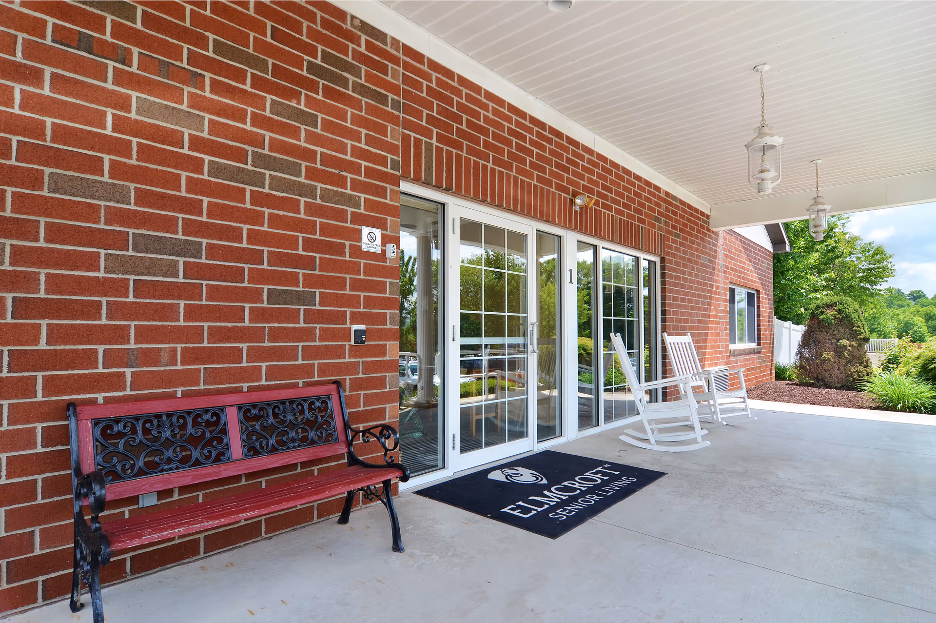 Covered brick entryway with a red bench, white rocking chairs, and glass sliding doors with a doormat.