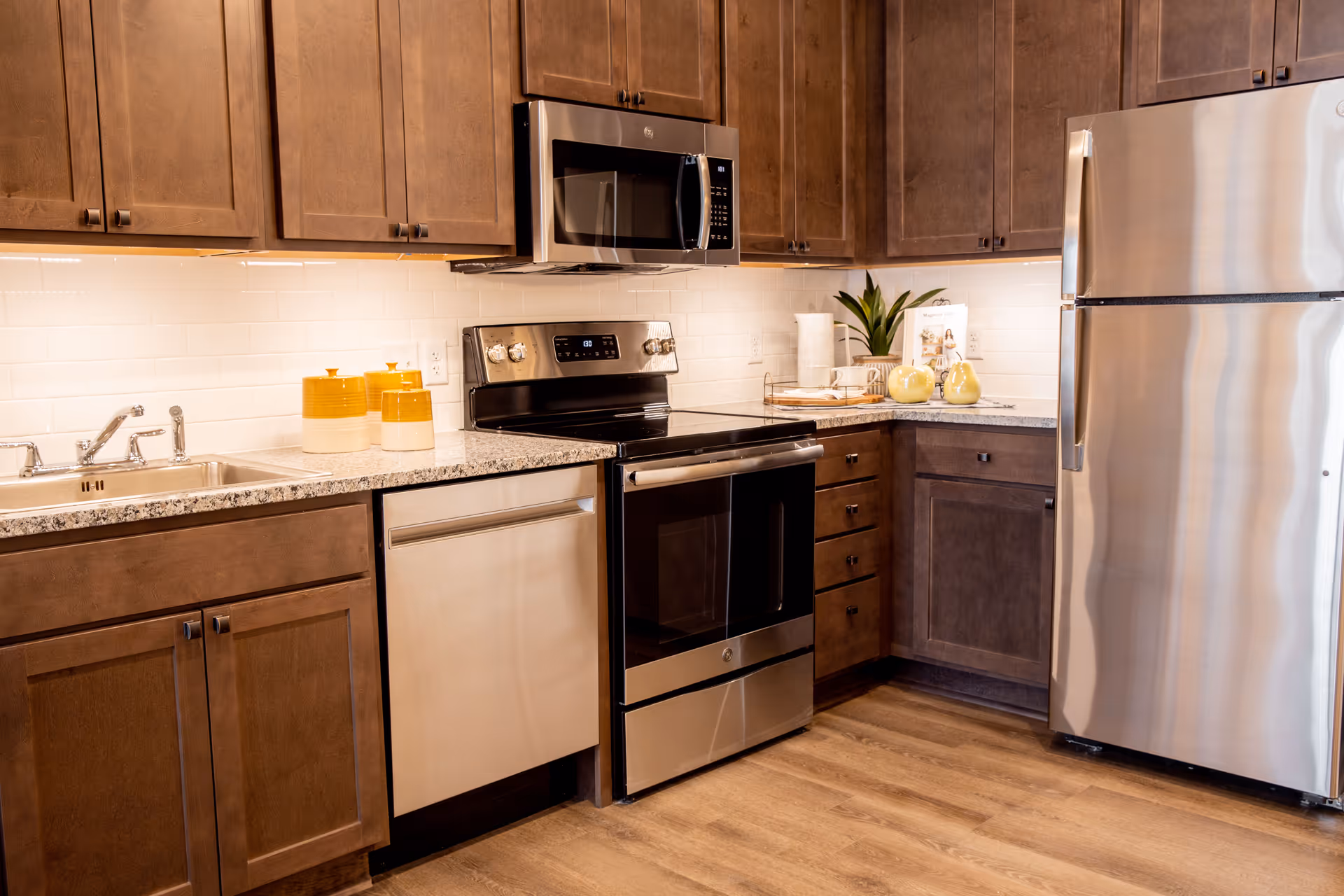Modern kitchen with wooden cabinets, stainless steel appliances including a refrigerator, oven, microwave, and dishwasher. The countertop is granite with a sink and decorative items such as yellow containers and a small plant.