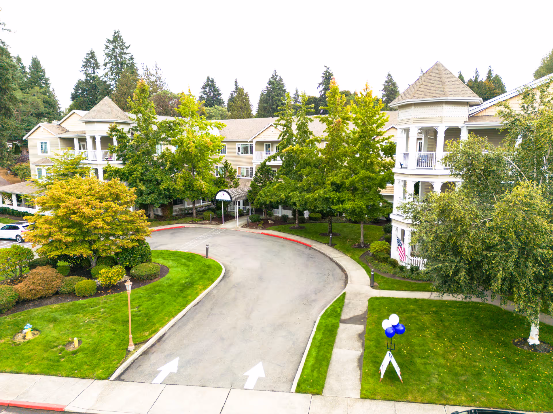 Front aerial view of a multi-story senior living building with wraparound balconies, a circular driveway, and landscaped lawns and trees.