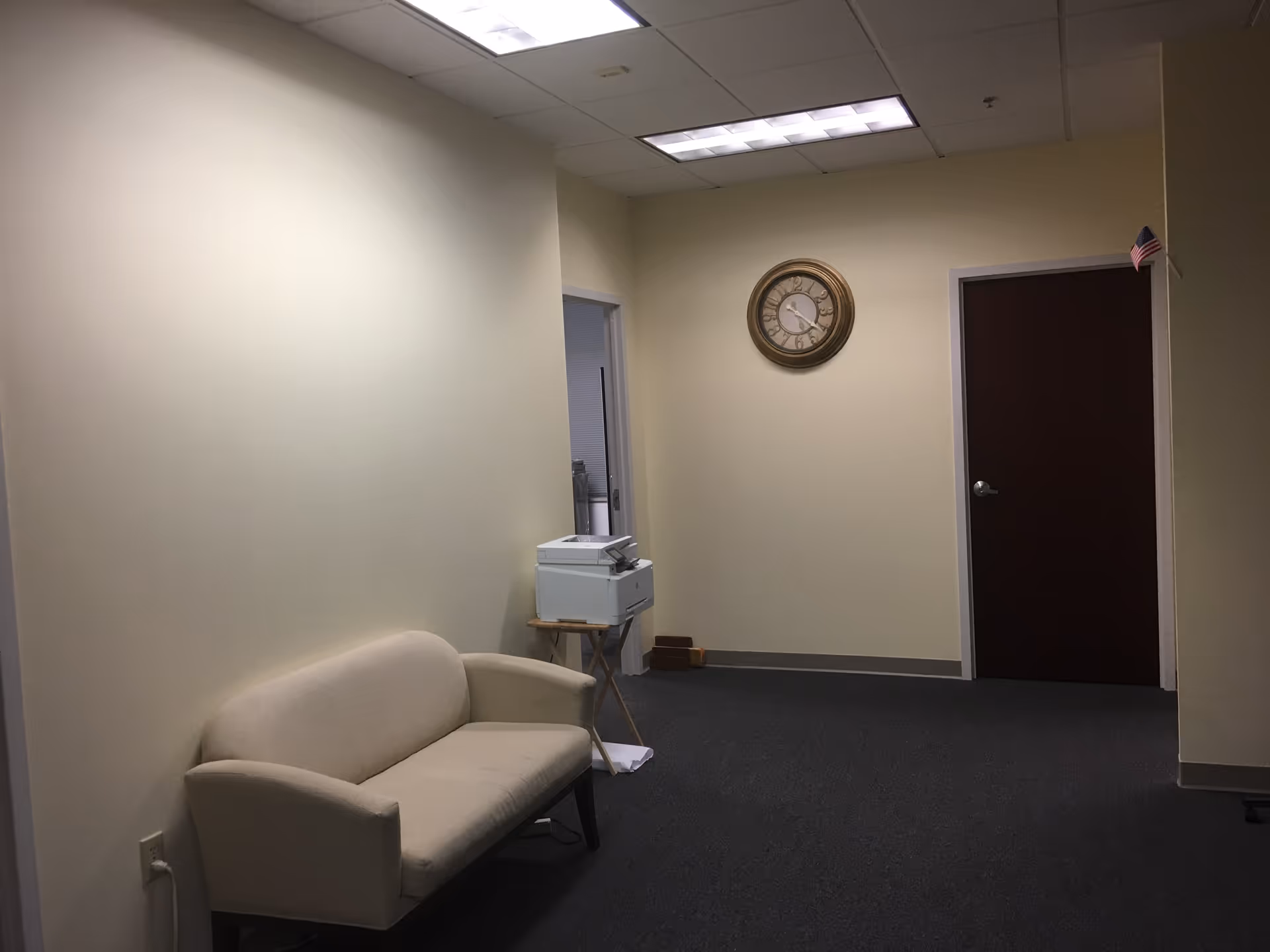 Plain beige waiting area along a hallway with a sofa, printer, clock, and door.