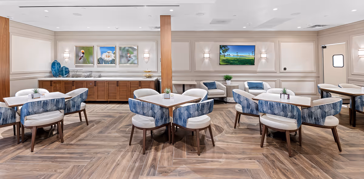 A bright and modern dining area with multiple square tables surrounded by blue and white cushioned chairs. The room features wood-patterned flooring, light-colored walls with decorative molding, and wall-mounted lights. On the back wall, there are three framed pictures of birds and flowers, a flat-screen TV displaying a golf course scene, and a wooden cabinet with decorative blue vases on top.