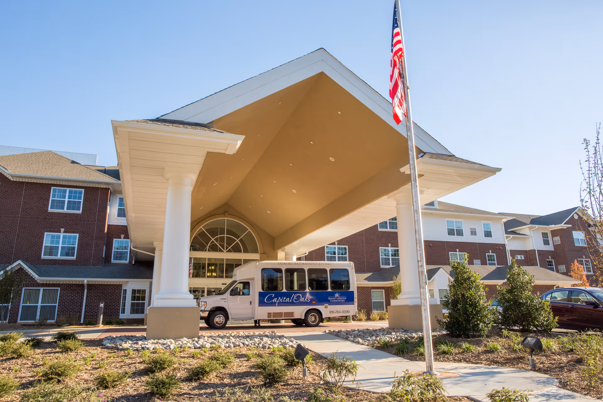 Front entrance of a brick retirement facility with a large covered porte-cochère, shuttle van, American flag, and landscaped driveway.