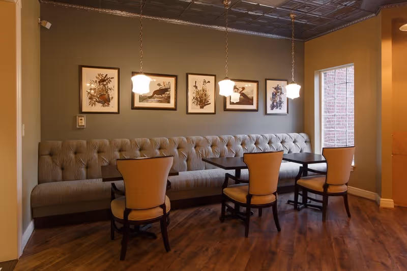 A cozy dining area with a long tufted bench along the wall, four wooden chairs with cushioned seats, two dark wooden tables, and three pendant lights hanging from a decorative ceiling. Five framed botanical prints are displayed on the wall above the bench, and a large window with a view of a brick wall lets in natural light.