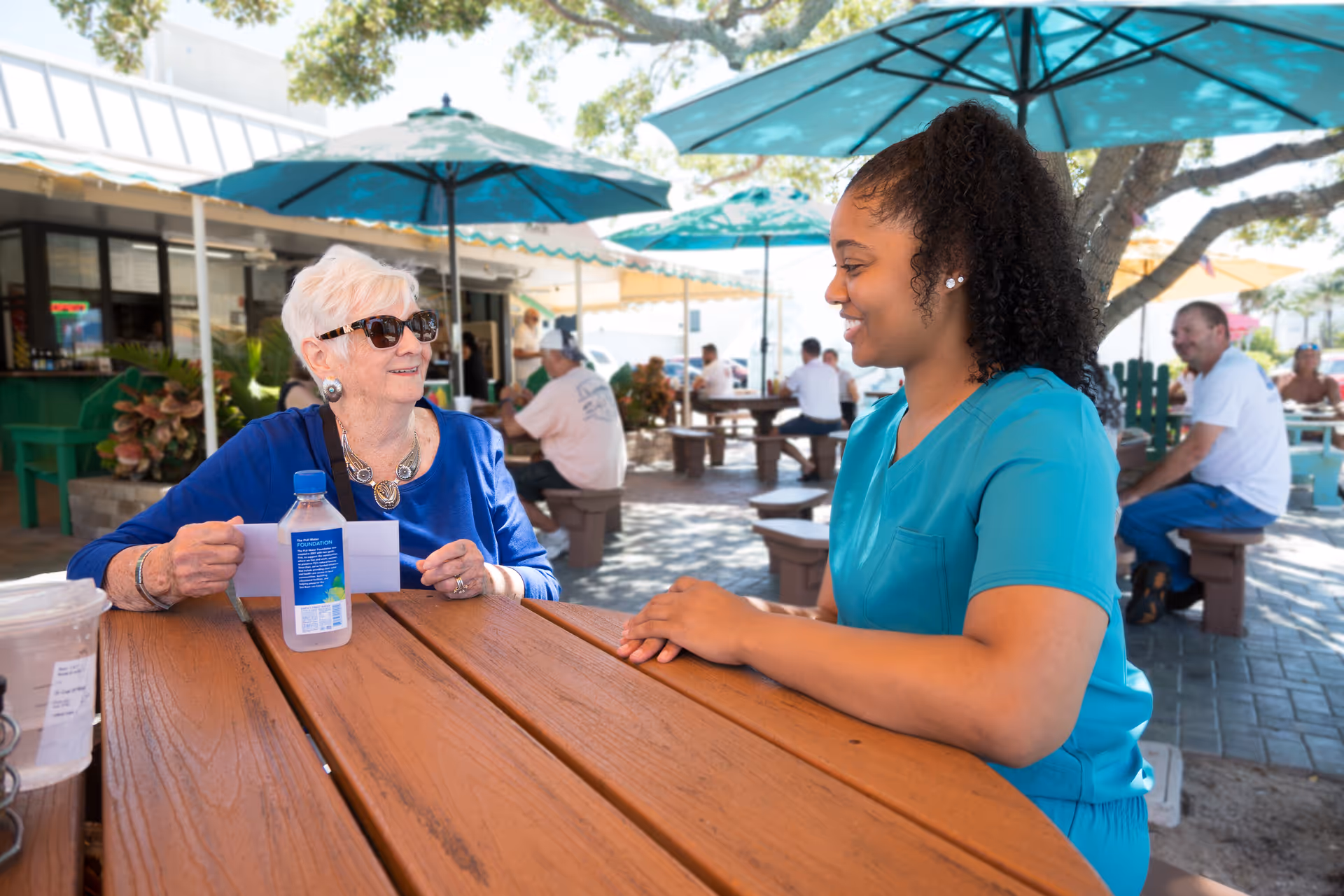 An elderly resident and caregiver chat at a wooden outdoor table under shade umbrellas.