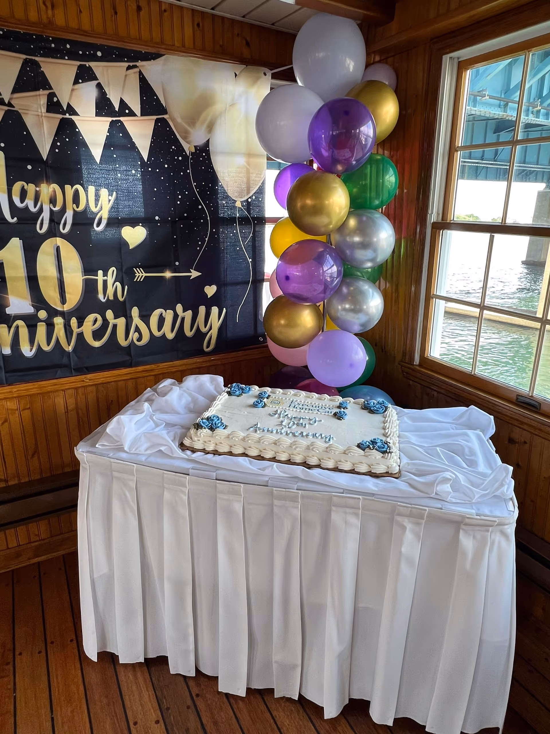 Indoor celebration setup with an anniversary cake, banner, and colorful balloons by a window.
