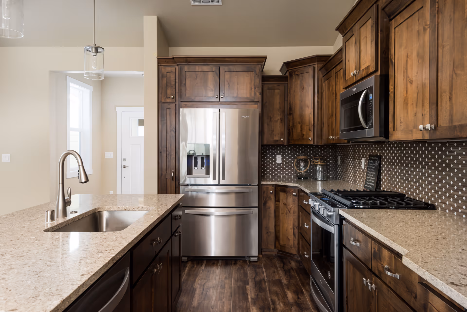 Modern kitchen with wooden cabinets, a stainless steel refrigerator, microwave, and stove. The kitchen features a large island with a sink and a sleek faucet, pendant lights hanging above, and a patterned backsplash.