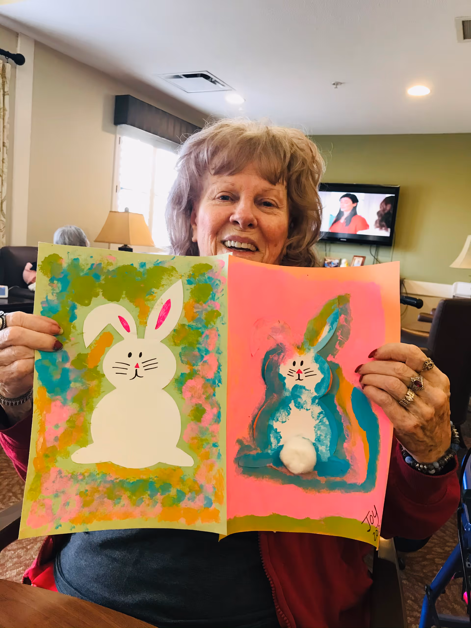 An elderly woman sitting indoors at Green Country Village holding up two colorful handmade Easter bunny art pieces, smiling. The room has a green wall, a TV in the background, and a lamp on a side table.