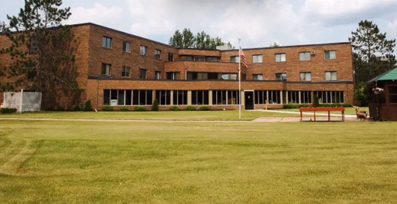 Brick senior living facility facade with green lawn and flagpole in front.