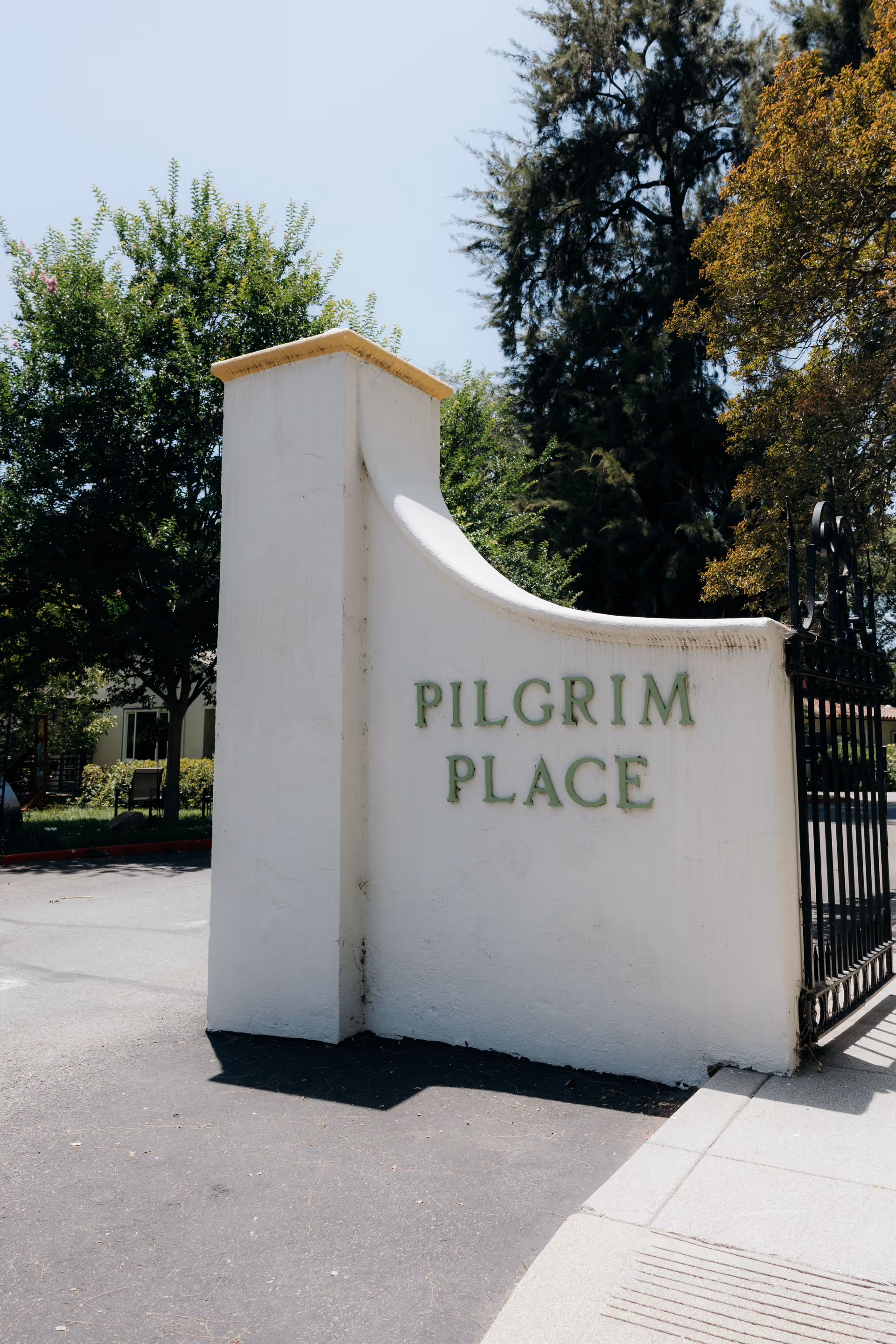 Entrance sign reading 'Pilgrim Place' on a white stucco wall next to a wrought-iron gate with trees in the background.